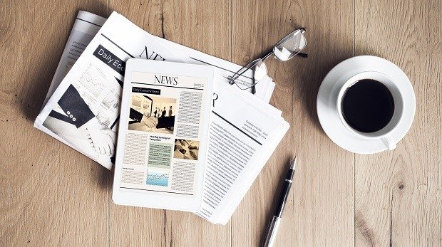 Tageszeitung und Tablet auf einem Holztisch. Daneben gefüllte Kaffeetasse und Kugelschreiber
