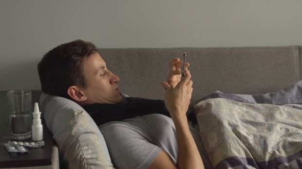 Mann liegt krank auf dem Sofa und bedient sein Smartphone