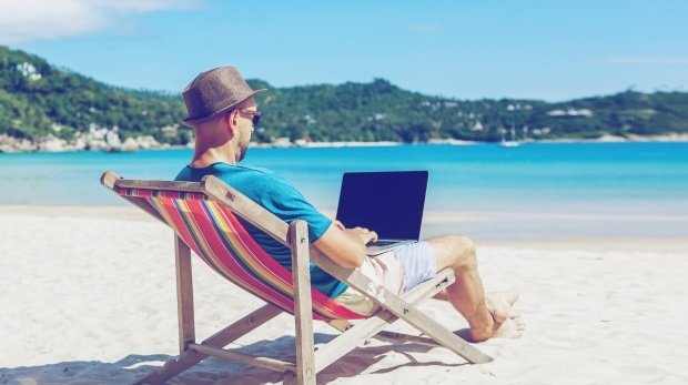 Mann sitzt mit Laptop am Strand im Liegestuhl