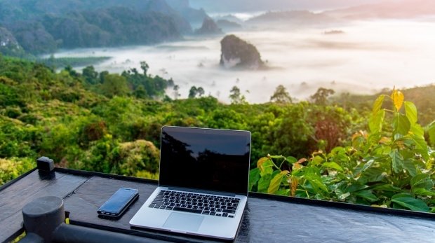 Ein Laptop steht auf einer Terasse mit einem Blick ins Grüne und auf die Berge