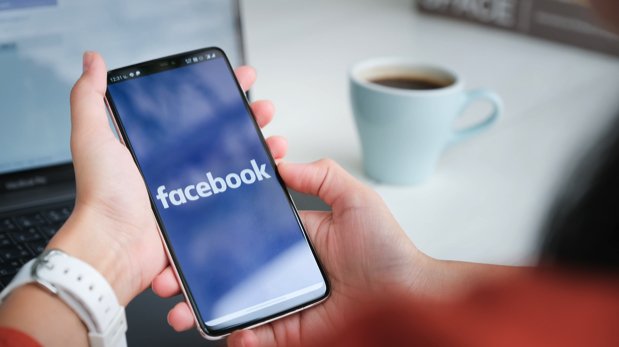 Smartphone mit Startsymbol der Facebook-App wird in der Hand gehalten. Notebook und Kaffeetasse im Hintergrund.