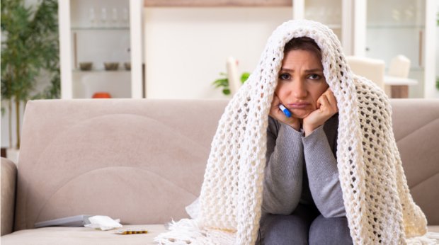 Eine Frau sitzt mit Decke und Fieberthermometer zu Hause auf dem Sofa