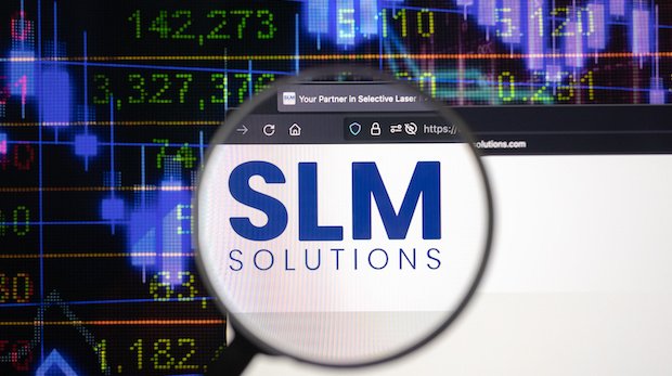 Logo SLM Solutions mit Lupe vor Hintergrund mit Börsenkursen