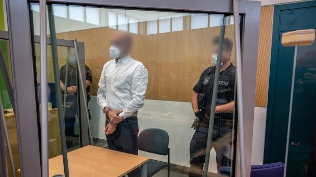 Der Angeklagte steht zwischen Justizbeamten hinter Sicherheitsglas im Landgericht Trier.