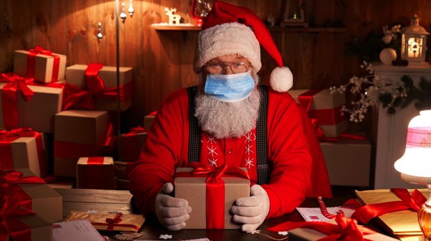Weihnachtsmann mit Mund-Nasen-Bedeckung am Tisch mit Geschenken