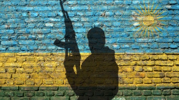 Schatten eines bewaffneten Mannes vor Wand, die in Farben der Nationalflagge Ruandas bemalt ist.