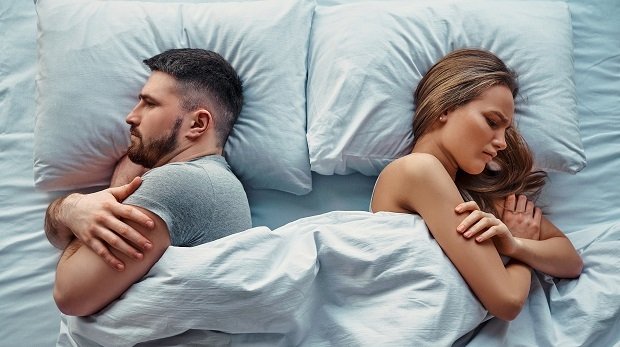 Ein junges Paar im Bett, voneinander abgewendet und mit verschränkten Armen.
