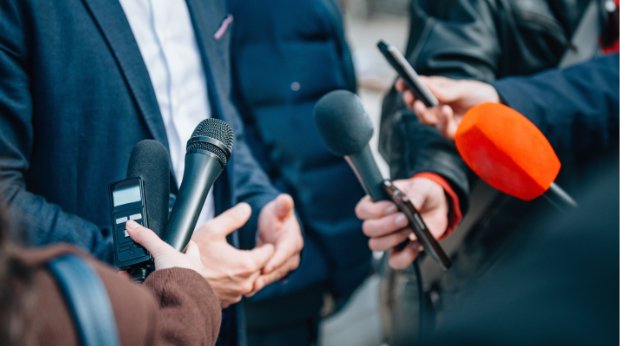 Ein Mann im Anzug wird von der Presse mit Mikrofonen interviewt