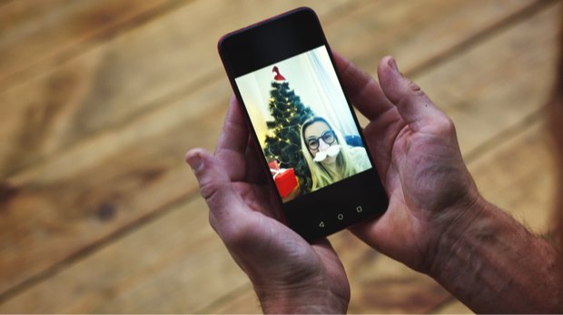 Hände halten Smartphone, auf dem ein Videocall mit einer Frau vor dem Weihnachtsbaum abgehalten wird.