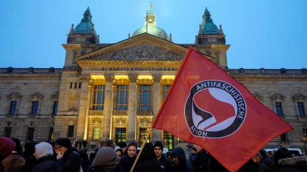 Teilnehmer einer linken Demonstration stehen mit einer Fahne mit der Aufschrift "Antifaschistische Aktion" vor dem BVerwG in Leipzig