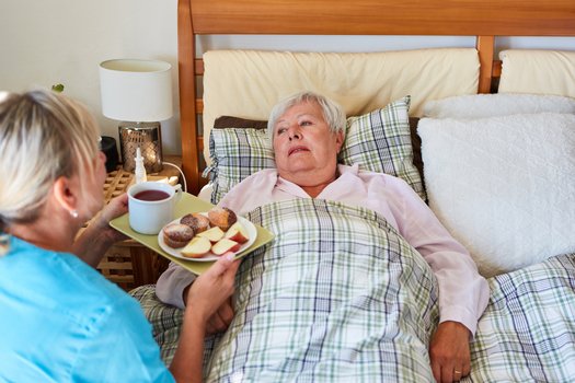 Eine Pflegekraft serviert einer alten Dame Essen ans Bett (Symbolbild)