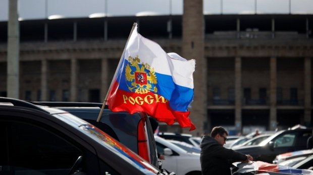 Flaggen mit den russischen Nationalfarben und dem russischen Staatswappen wehen an einem Auto auf dem Olympiaplatz vor dem Olympiastadion.